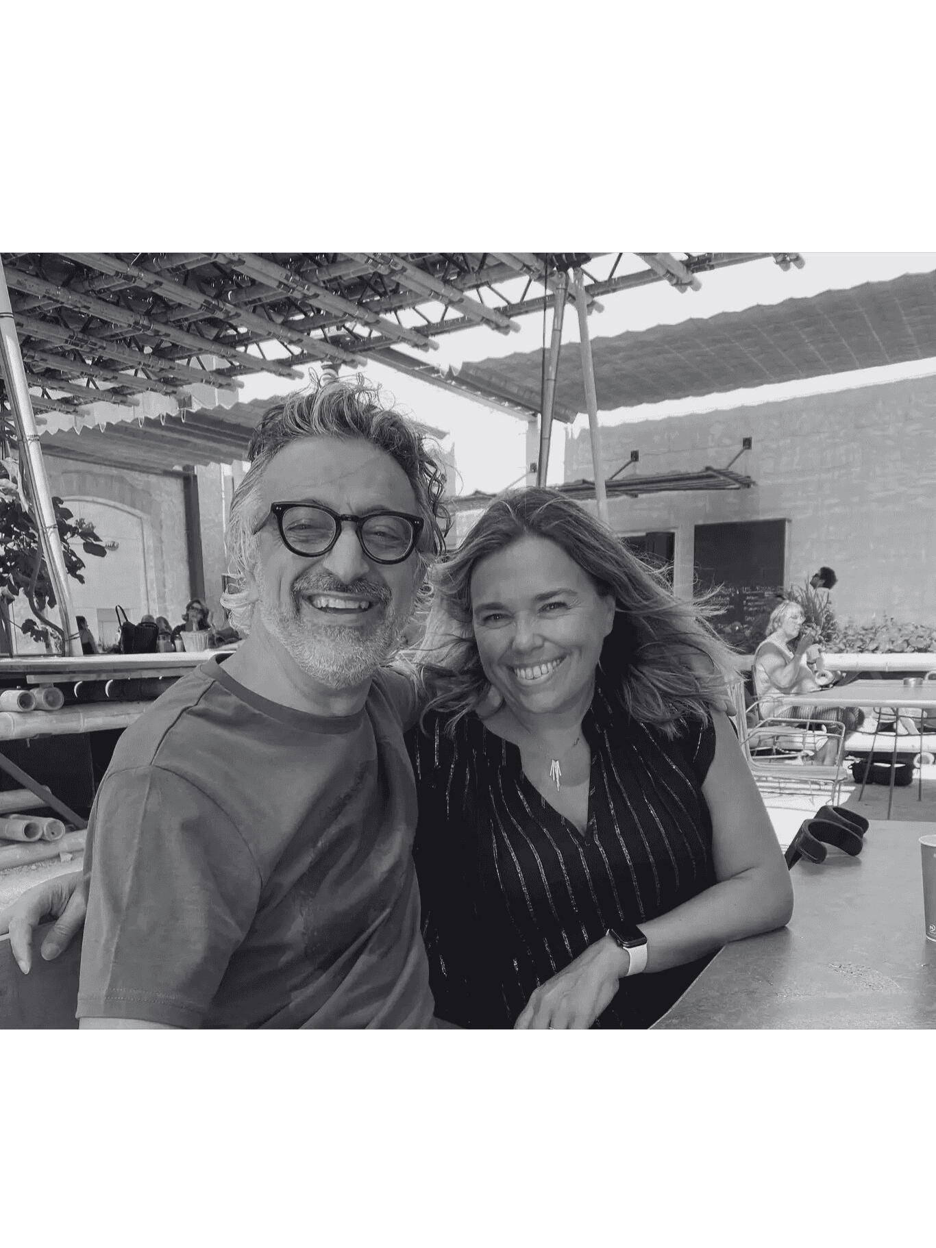 A smiling man and woman sit together at an outdoor cafe, enjoying a sunny day.